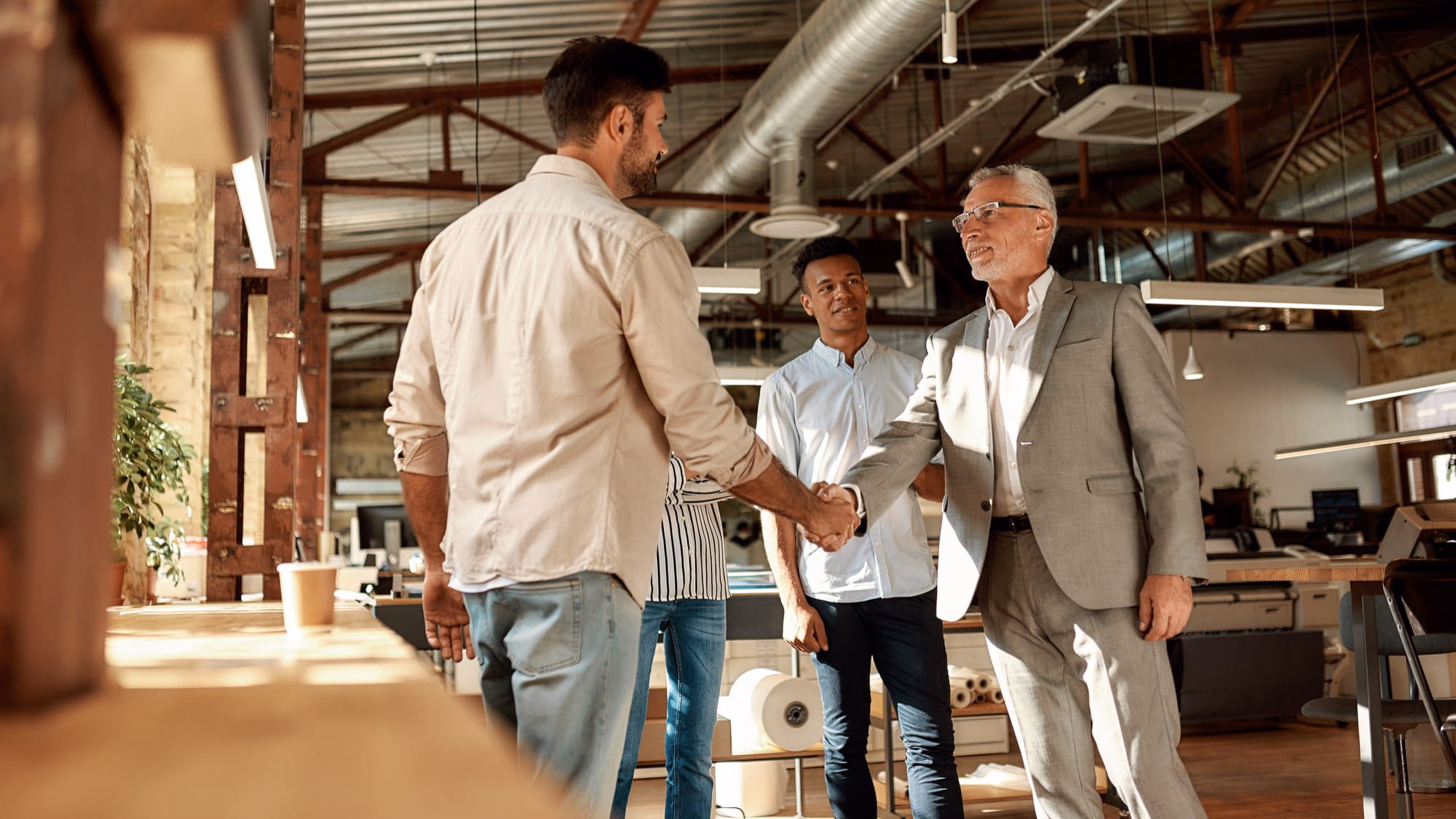 Two cheerful men shaking hands and smiling while standing with coworkers in the modern office